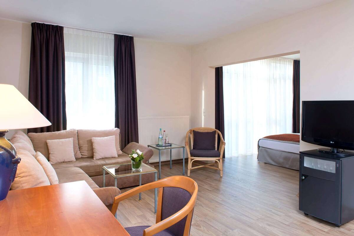 A modern hotel room with a sofa, a coffee table, a TV, and a dining area.