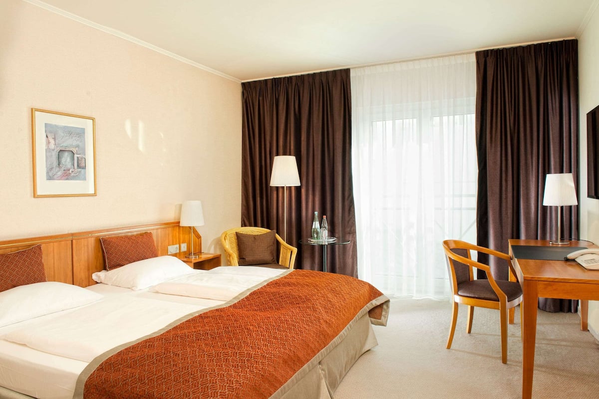 A hotel room with a bed, two chairs, a desk, a lamp, and a painting on the wall.