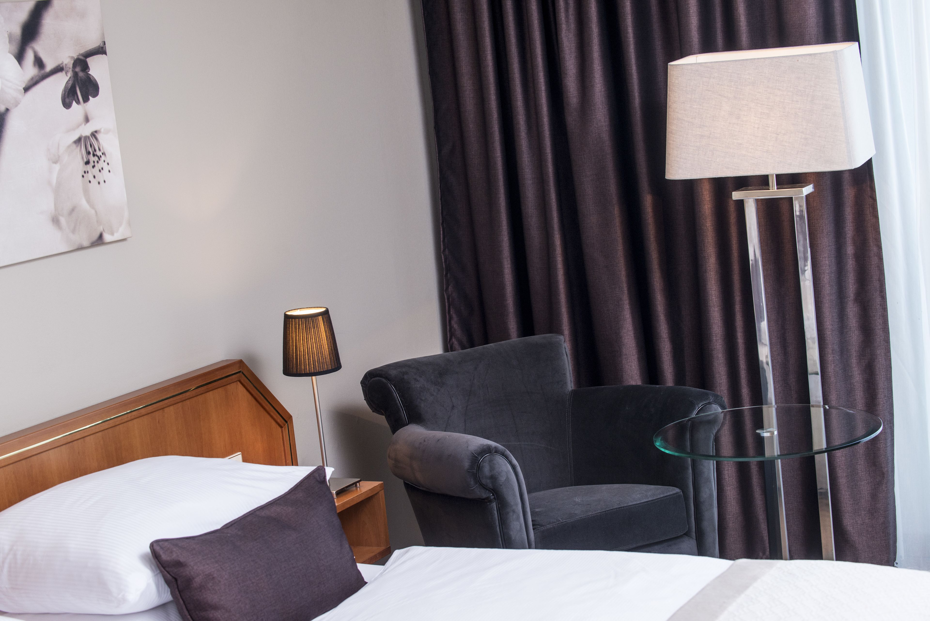 Habitación de hotel con cama, una silla, una mesita de noche con lámpara y una mesita de vidrio con lámpara.