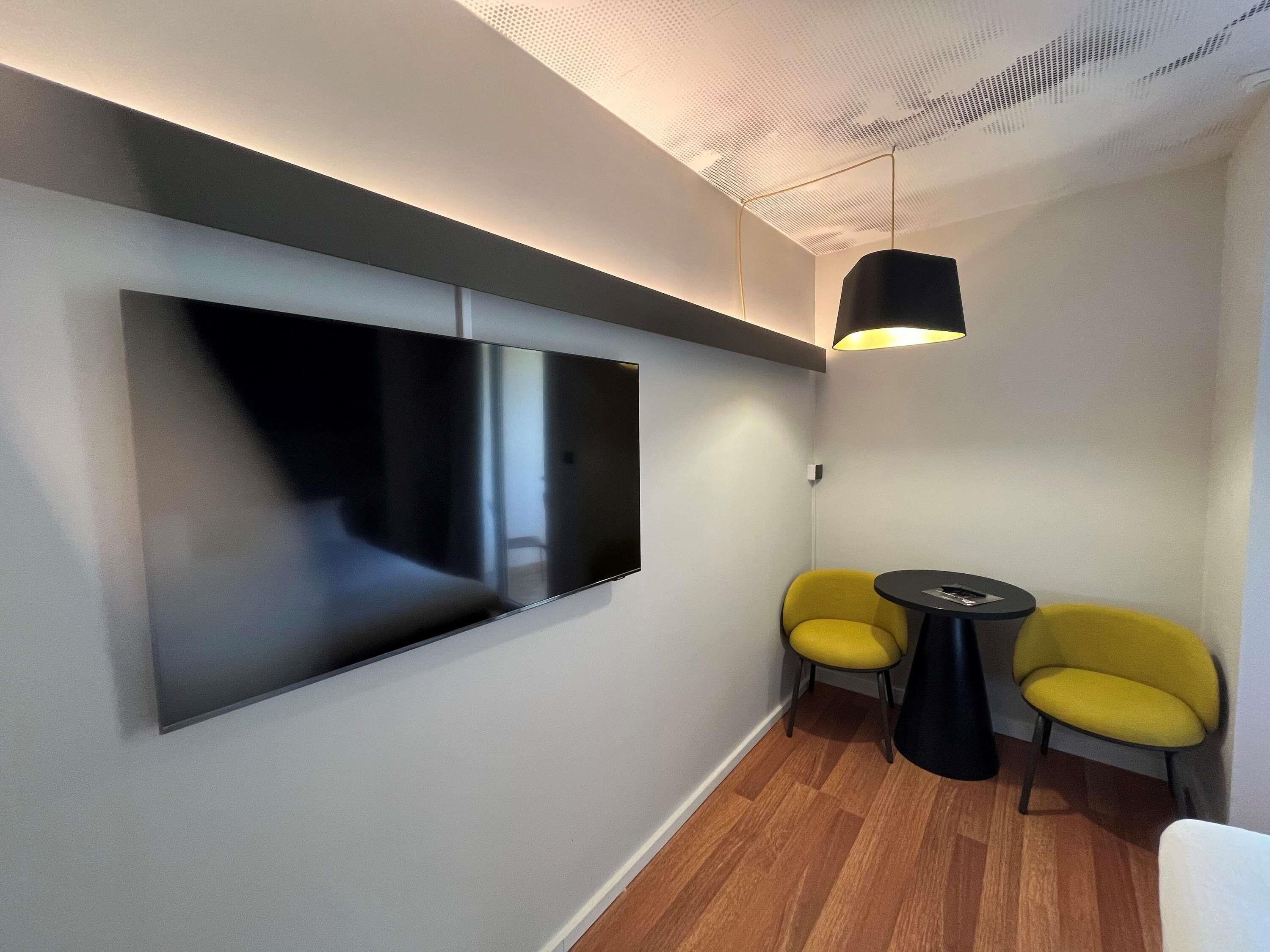 Een moderne kamer met een flatscreen televisie aan de muur, twee gele stoelen rond een klein zwart tafeltje, en een hanglamp.