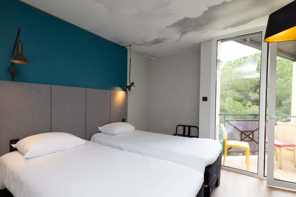 A modern hotel room with two beds, a balcony with outdoor seating, and a view of greenery.