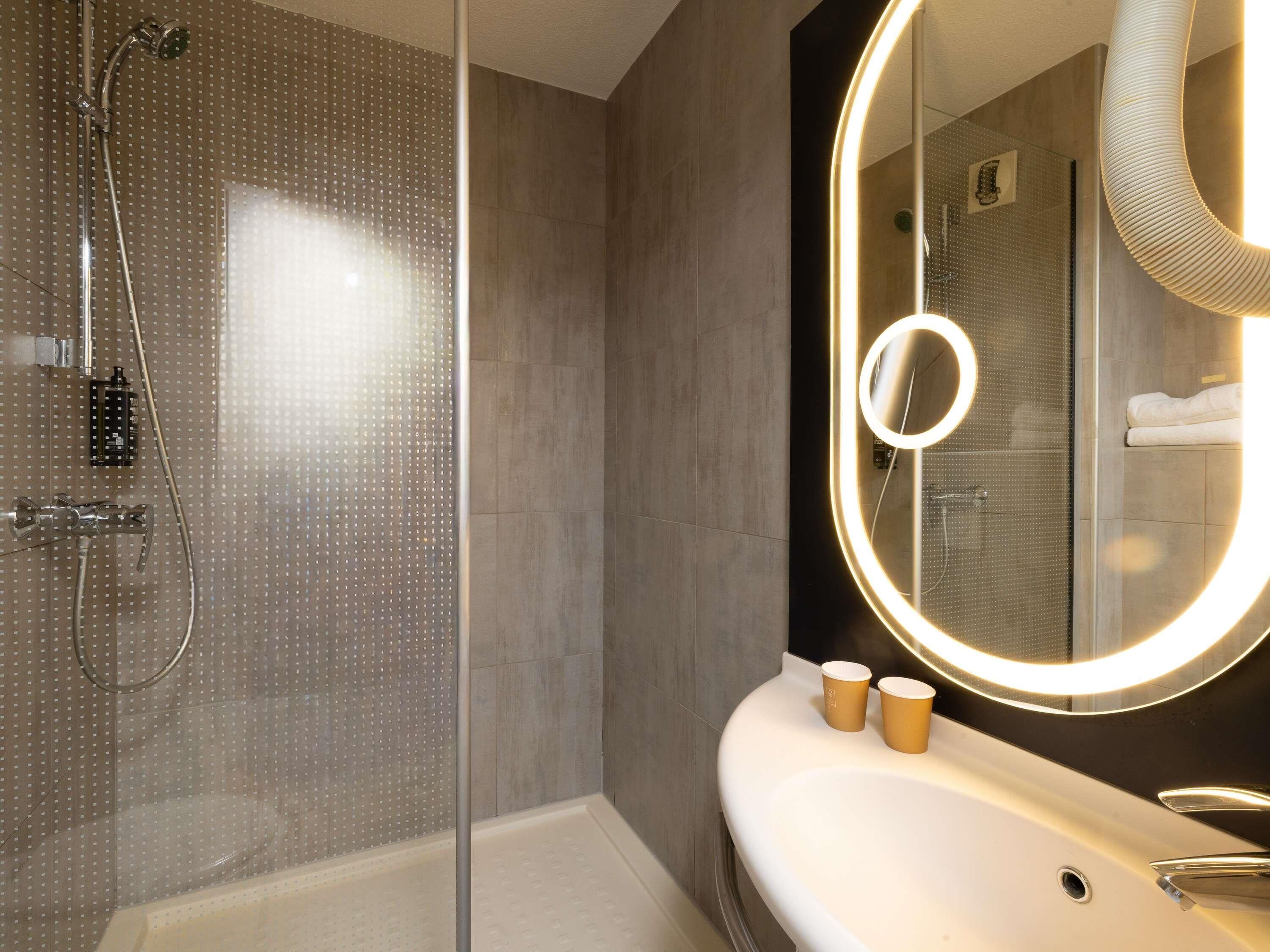 A modern bathroom with a glass-enclosed shower, a large mirror with a circular light, and a white sink with two cups.