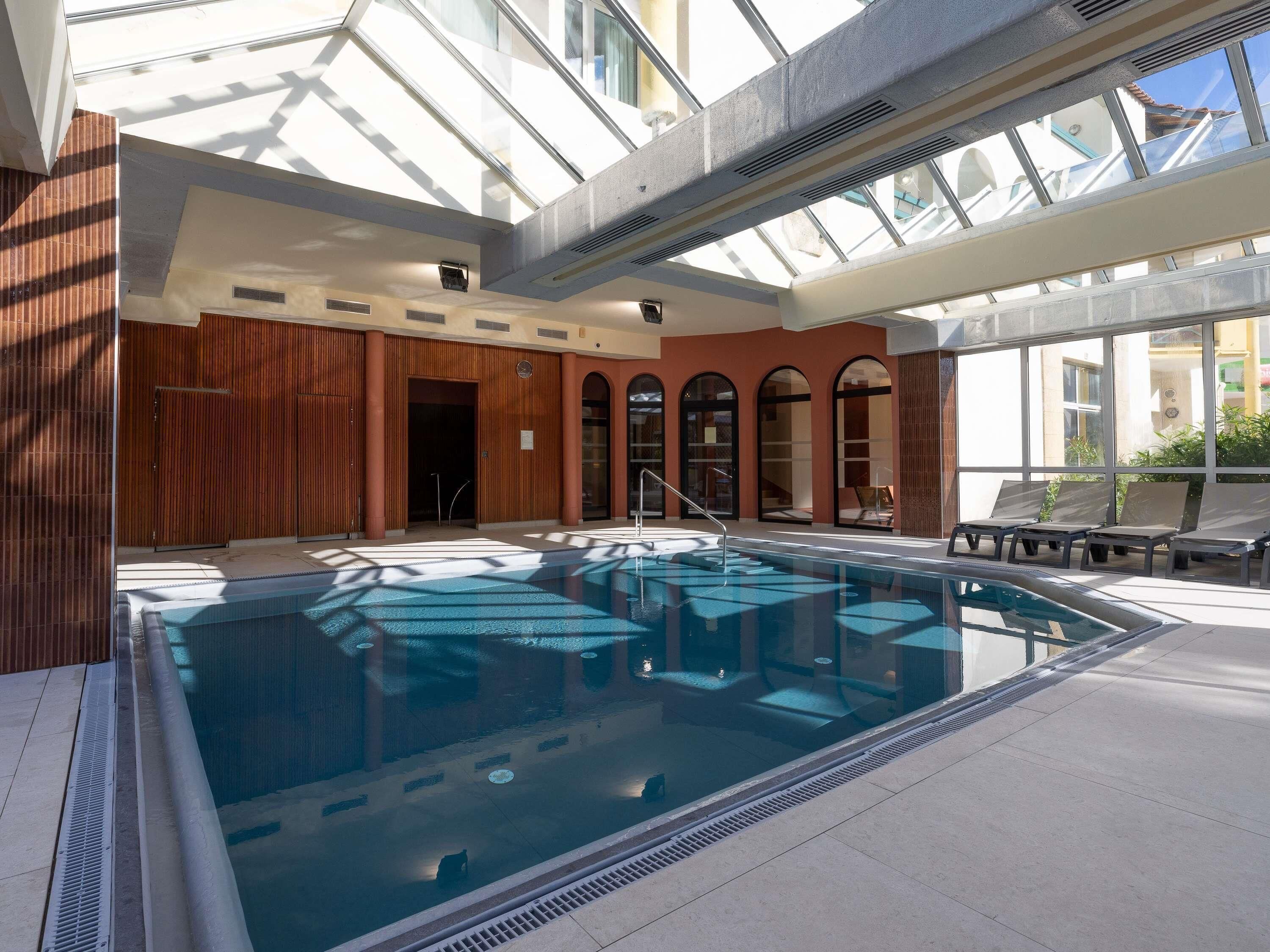 An indoor swimming pool with a glass ceiling, sunlit lounge area, and a view of the outside.
