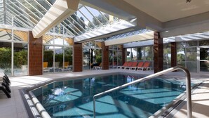 Ein überdachter Pool mit großen Glaswänden, ein sonnendurchfluteter Liegebereich und Blick auf Grünfläche.