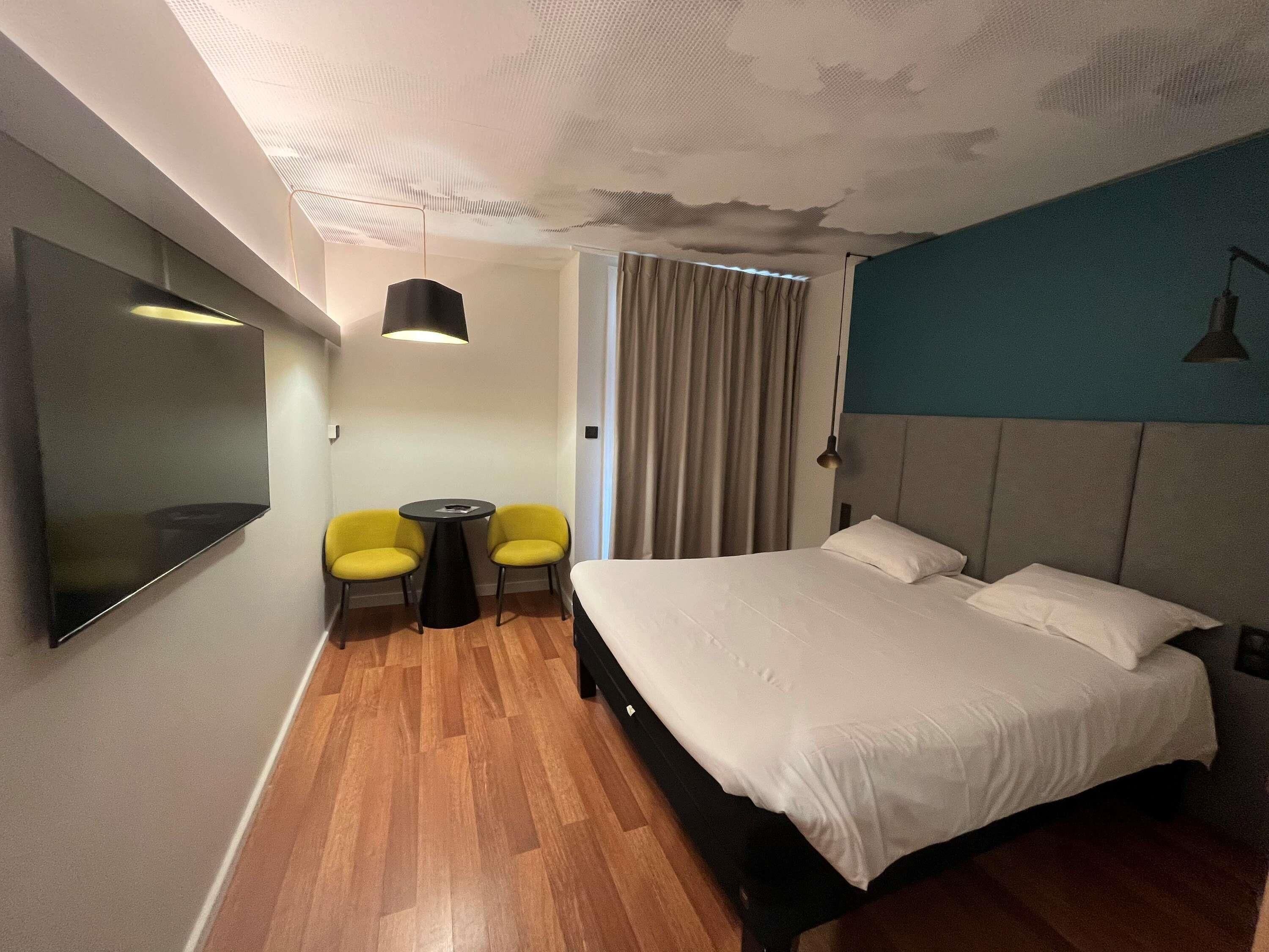 Une chambre d’hôtel avec un lit, une télévision, deux chaises jaunes, une petite table et un luminaire au plafond.