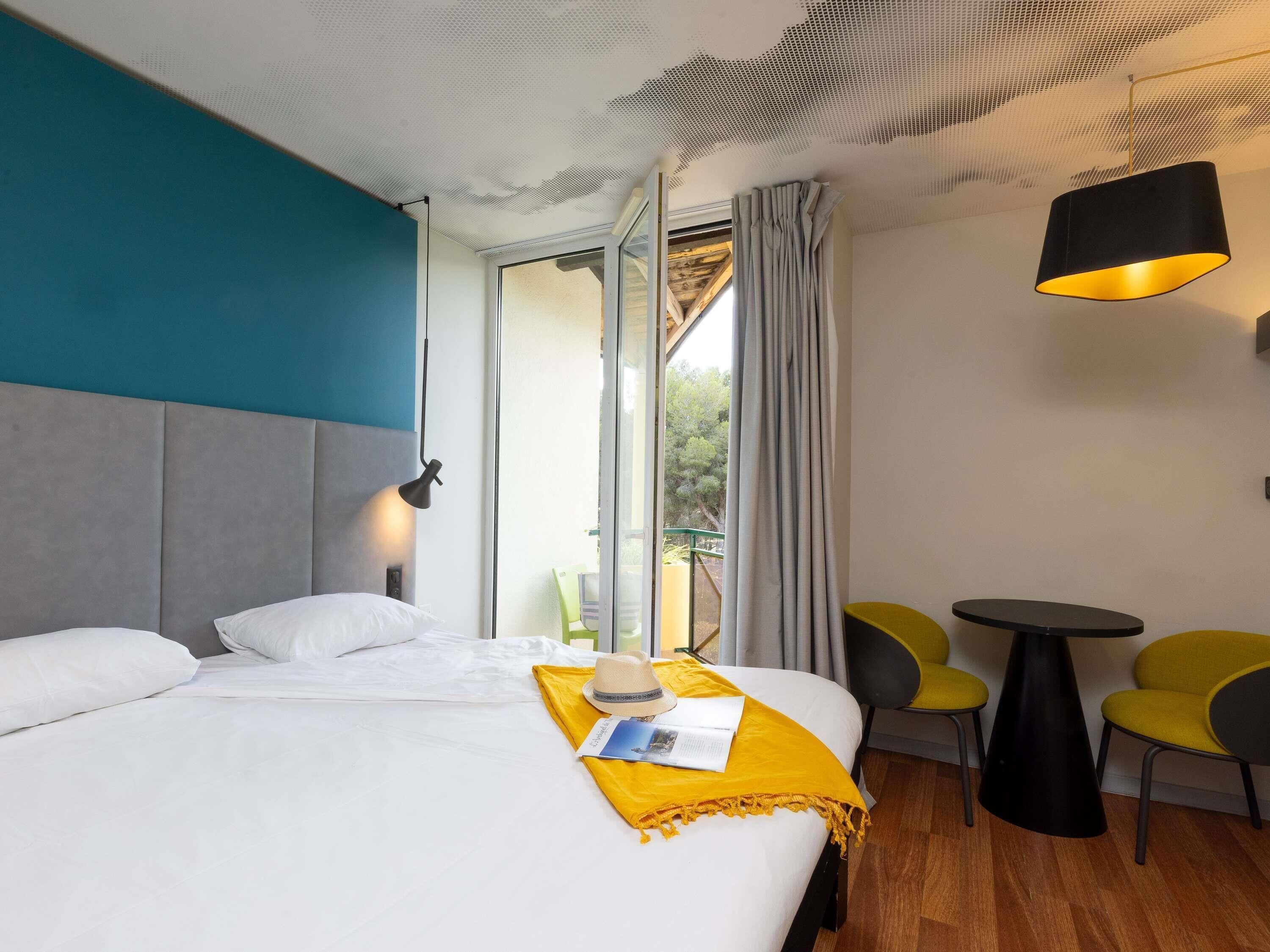 A hotel room with a bed, a small table, chairs, a ceiling light, and a view of the outdoors.
