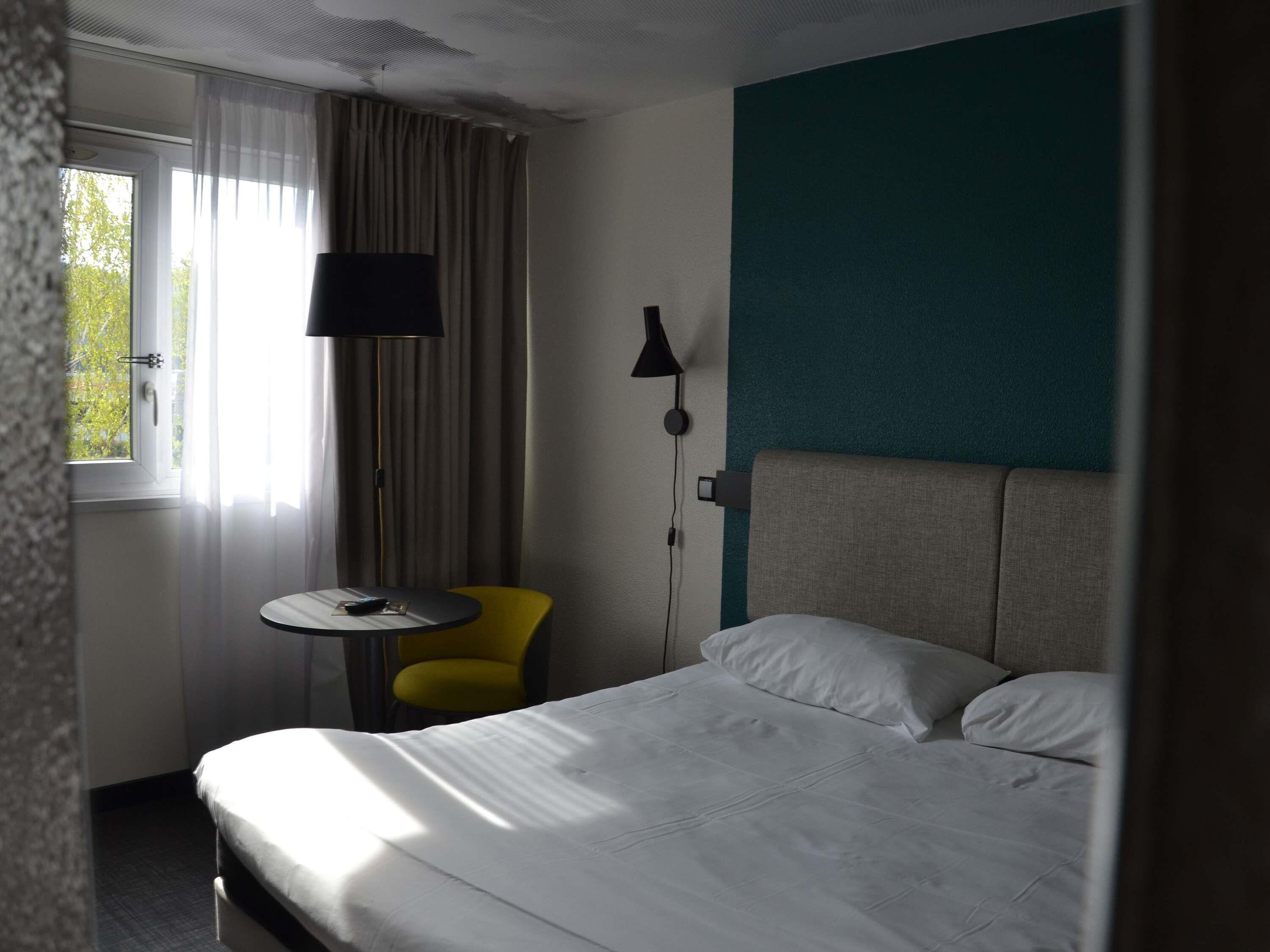 Habitación de hotel con cama, una mesita, una silla, una lámpara y ventana con cortinas.