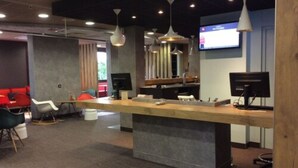 Un área de recepción moderna con un mostrador de madera, diversas opciones de asientos que incluyen un sofá rojo y sillas blancas, y una pantalla grande que muestra información.