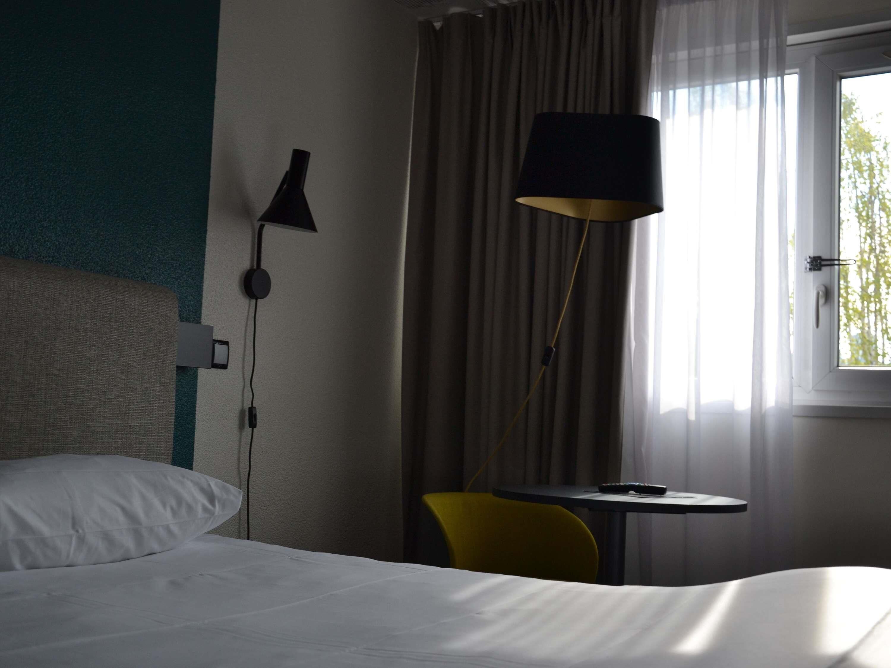 A hotel room with a bed, a desk, a lamp, and a window with curtains.