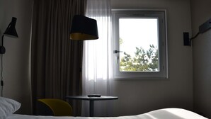 Habitación de hotel con cama, una mesita, una silla, una lámpara y ventana con vista a árboles.