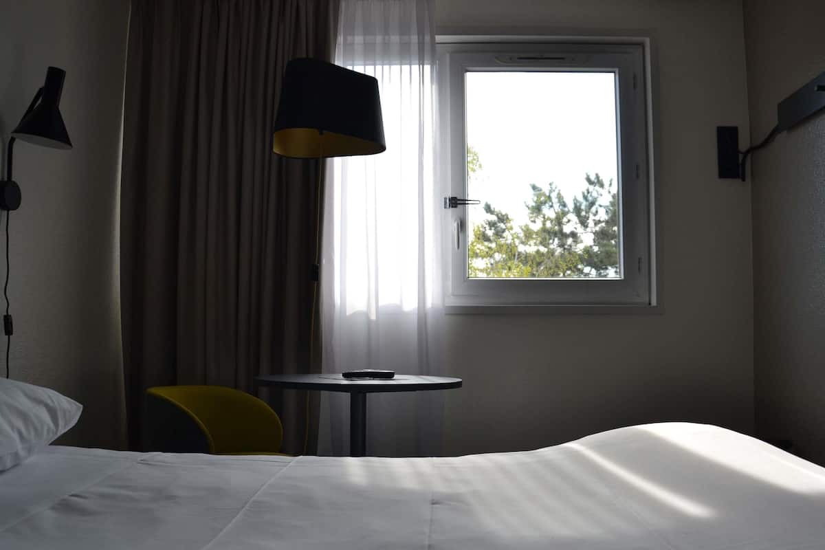 Habitación de hotel con cama, una mesita, una silla, una lámpara y ventana con vista a árboles.