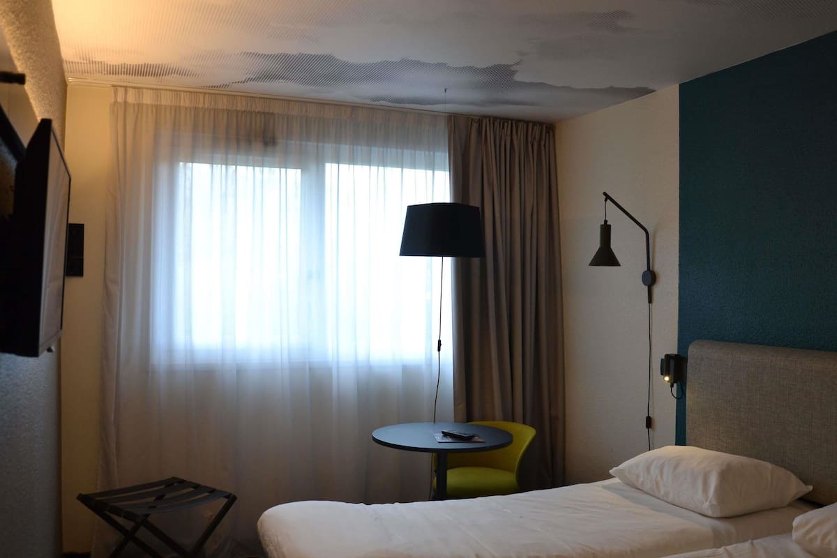 Una habitación de hotel con cama, una mesita, una silla, una lámpara y un ventanal con cortinas.