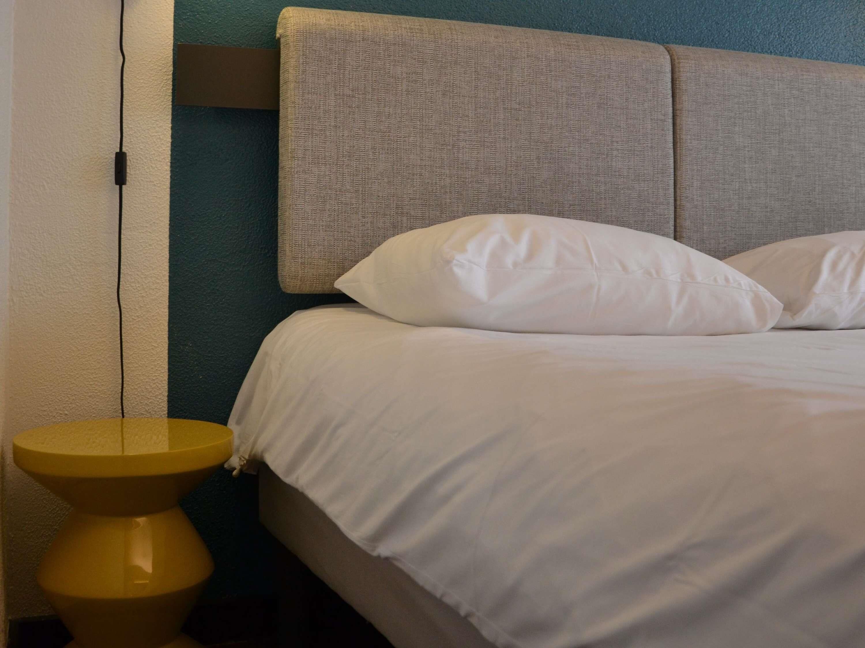 Uma cama com roupa branca, cabeceira e mesa de cabeceira com abajur.