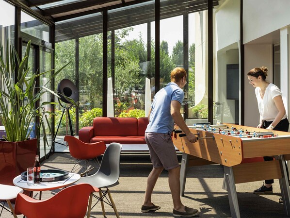 A modern indoor space with a foosball table, red seating, and large windows offering a view of greenery. - ibis Marne La Vallee Emerainville (Emerainville)