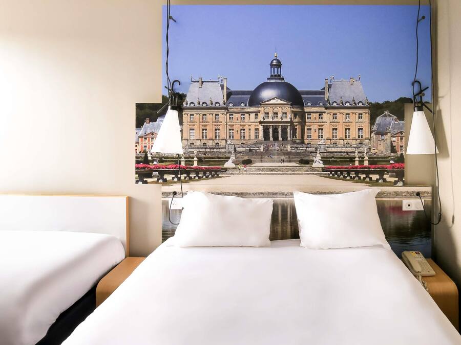 Ein Hotelzimmer mit einem Bett, zwei weiße Kissen, einem Nachttisch mit einer Lampe und einem an der Wand befestigten Telefon.