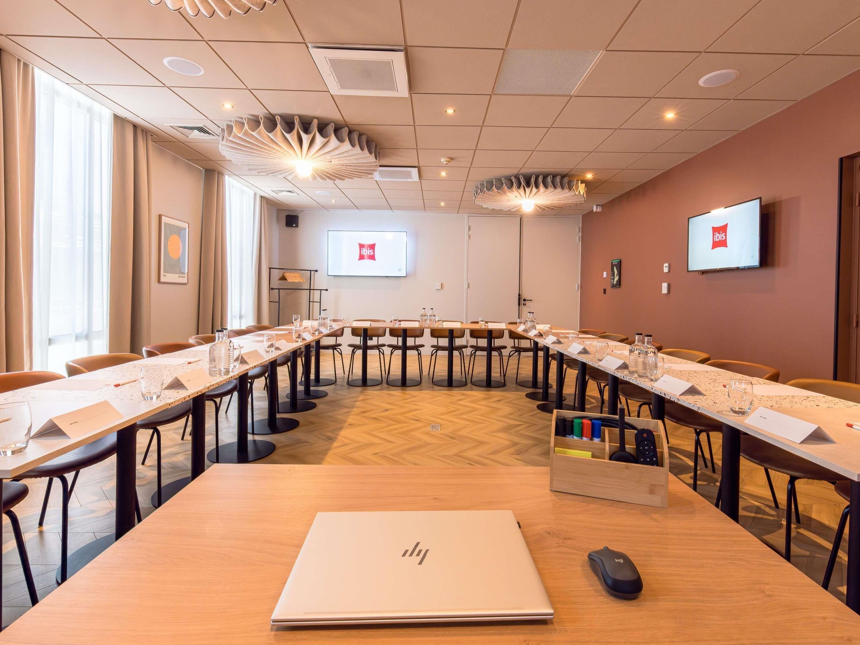 Un salón de conferencias con una larga mesa, sillas, una computadora portátil, un ratón y una pantalla de proyector que muestra el logotipo de ibis.