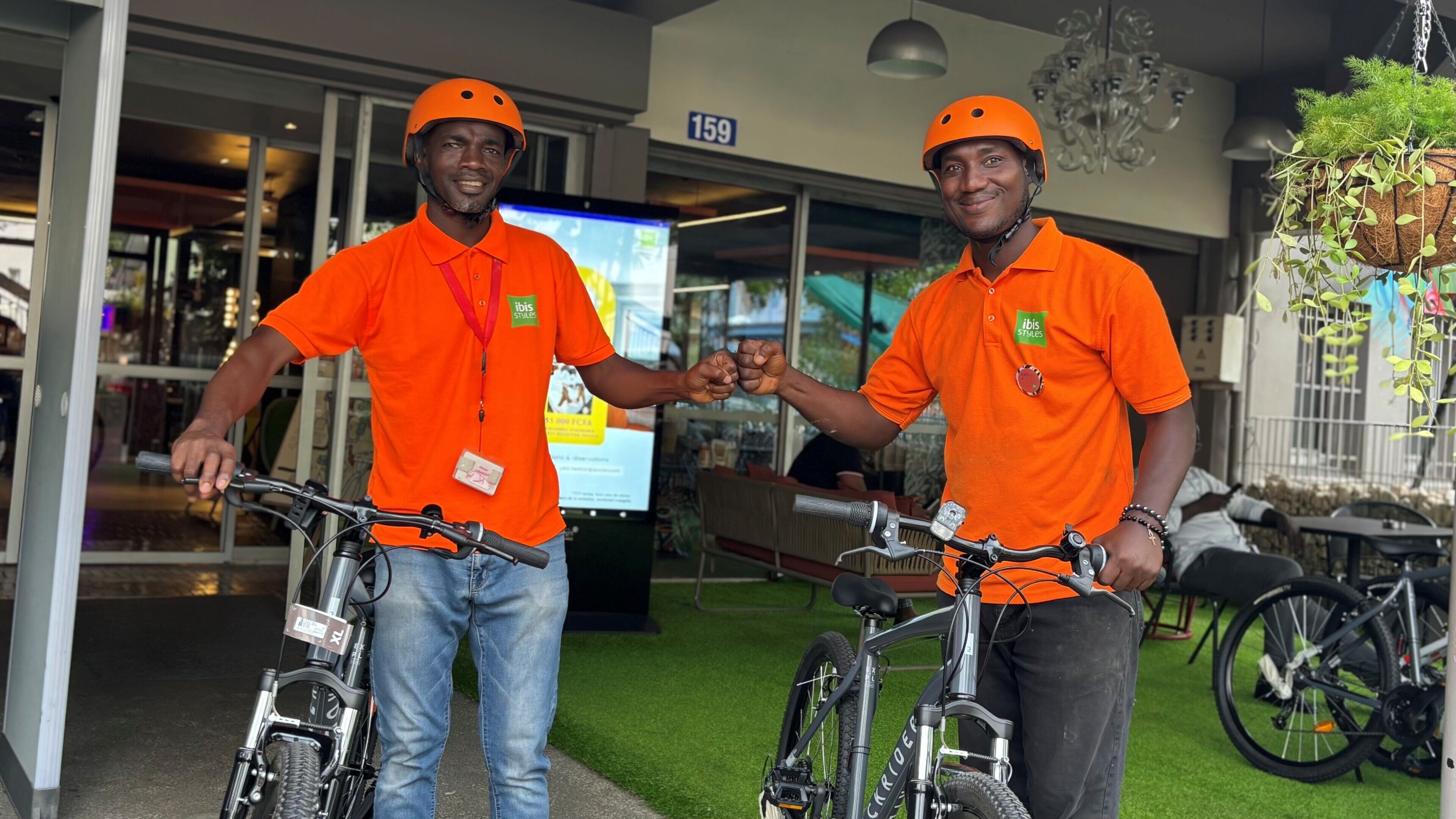 Two individuals in orange shirts with helmets, holding bicycles, standing in an indoor area with a green floor, a visible number '159', and a hanging plant.