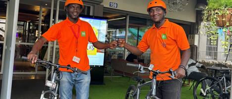 Deux personnes portant des maillots orange et des casques, tenant des vélos, se tiennent dans un espace intérieur doté d’un sol vert, où l’on peut voir le numéro « 159 » et une plante suspendue.