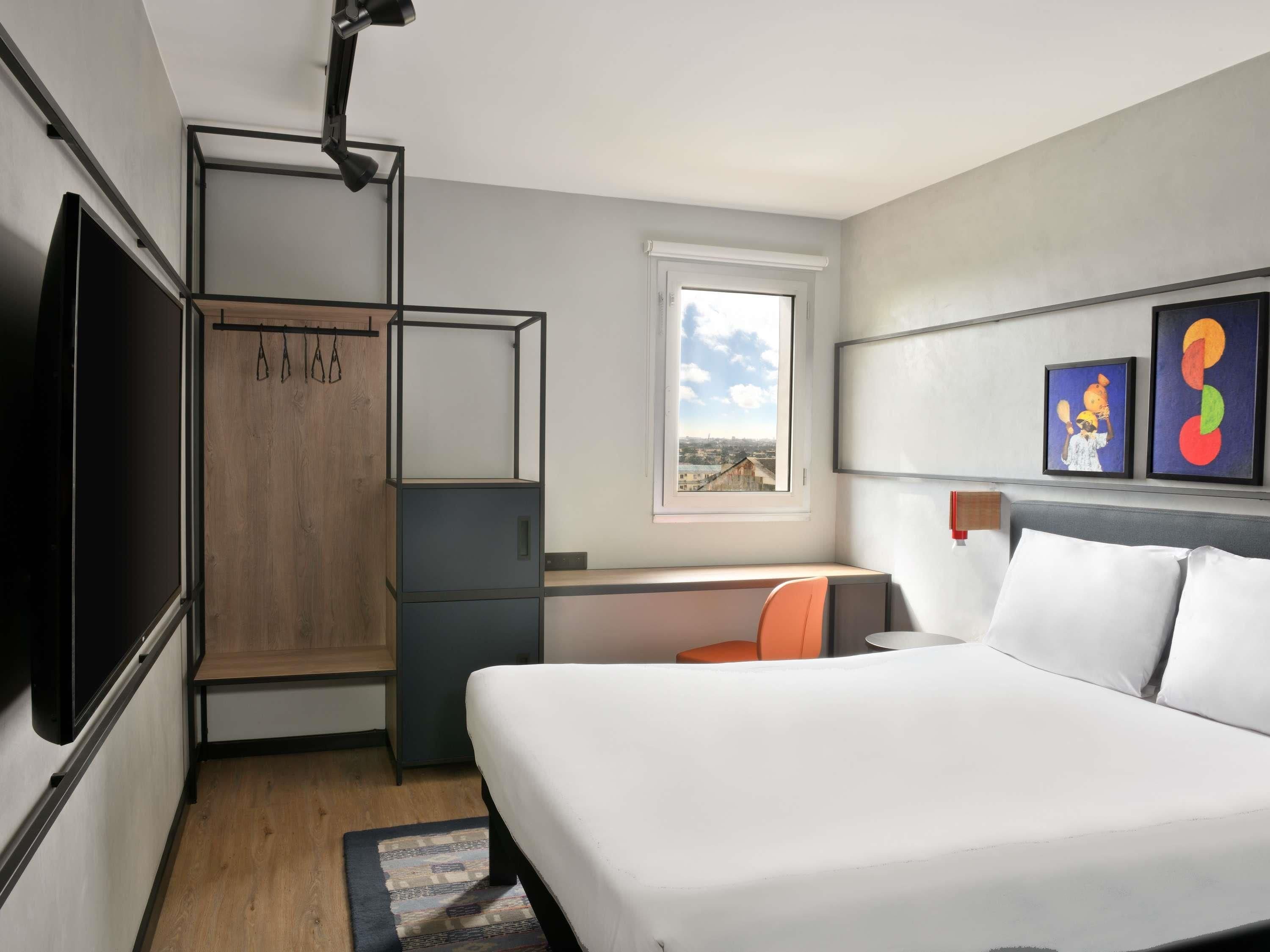 A modern hotel room with a bed, desk, and a view of the city through a window.