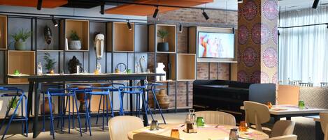 Un comedor moderno con una mezcla de sillas azules y beige, una mesa central con un mantel individual colorido y un televisor montado en la pared.