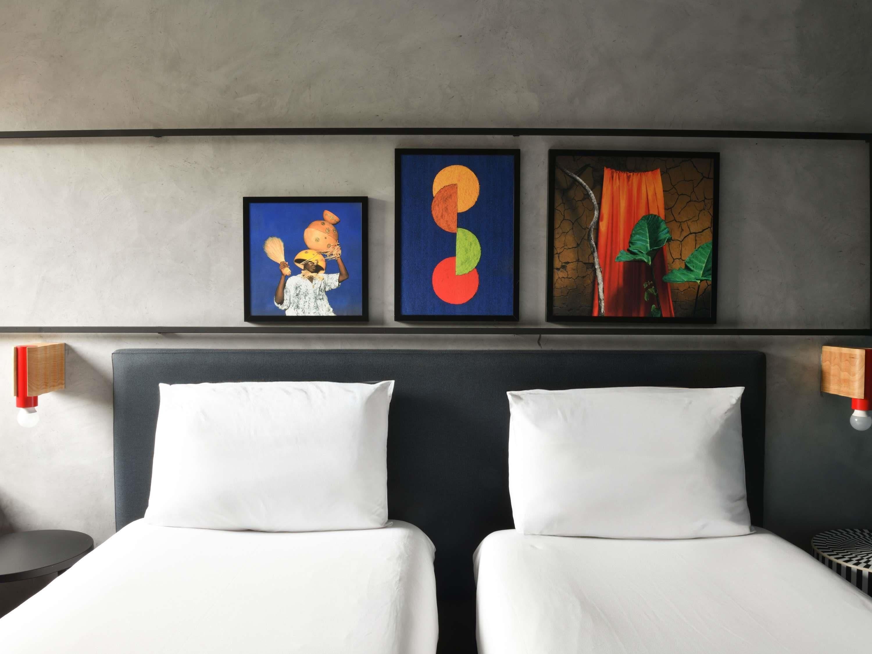 A hotel room with two beds, a headboard, and three framed artworks on the wall.