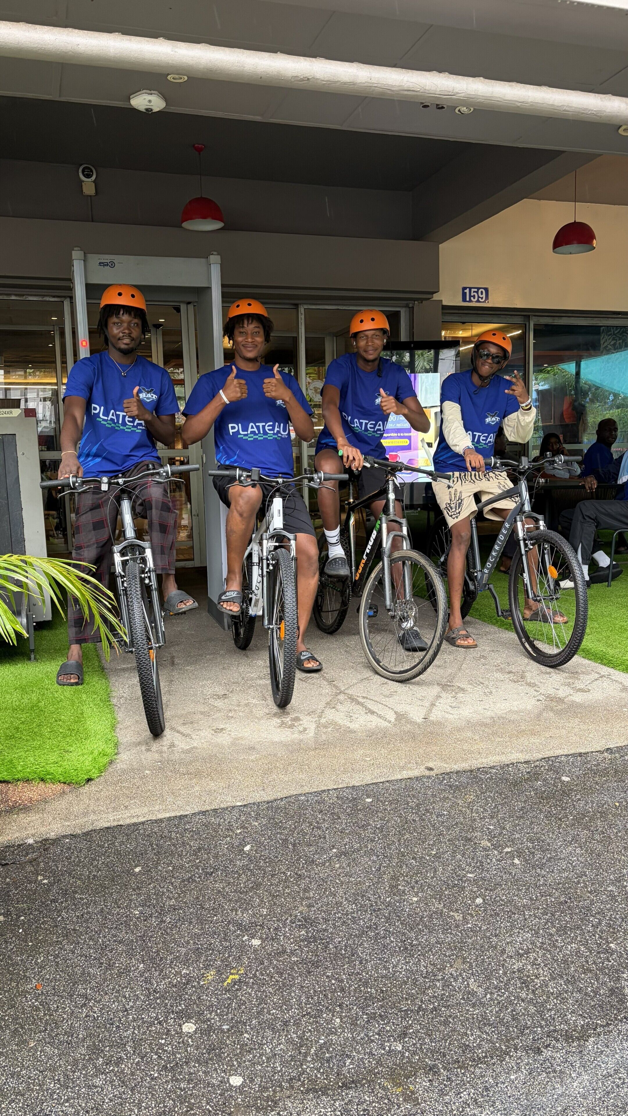 Quatre personnes à vélo se trouvent devant un bâtiment. Une enseigne portant le numéro « 159 » est placée au-dessus de l’entrée.
