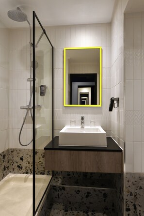 A bathroom with a glass-enclosed shower, a white sink with a black countertop, and a mirror with a yellow frame. - ibis Styles Abidjan Plateau (Abidjan)