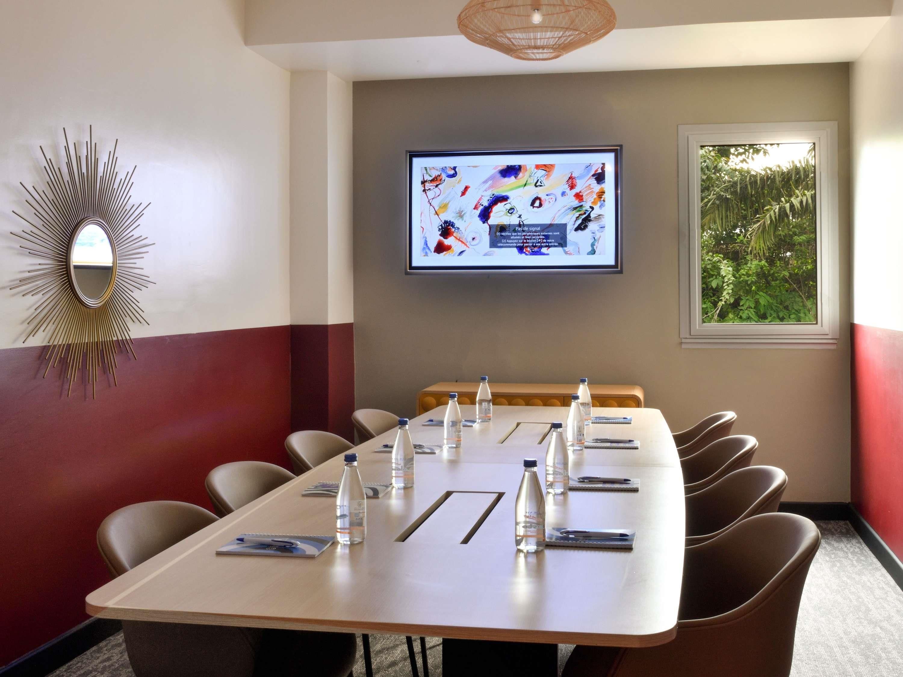 Un salón de conferencias con una mesa larga, sillas, botellas de agua y una pantalla de televisión que muestra una imagen.