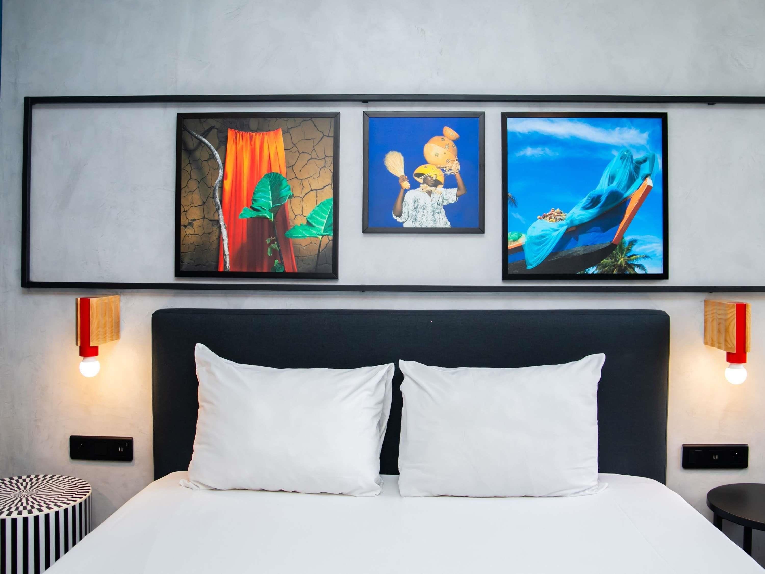 A hotel room with a bed, two pillows, a bedside table, a round side table, and three framed artworks on the wall.