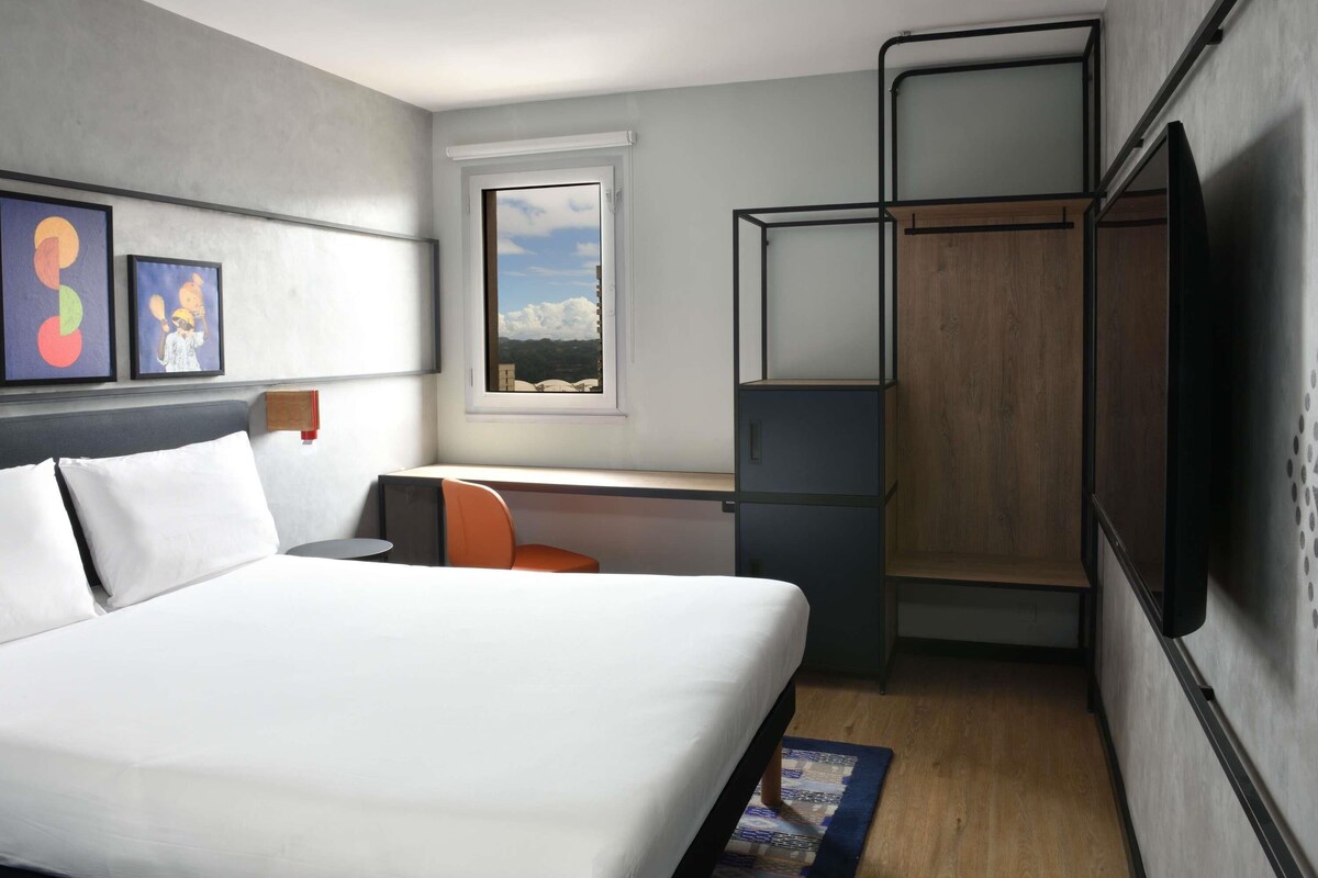 A modern hotel room with a large bed, a desk with a chair, a wardrobe, and a window with a view of the sky and clouds.