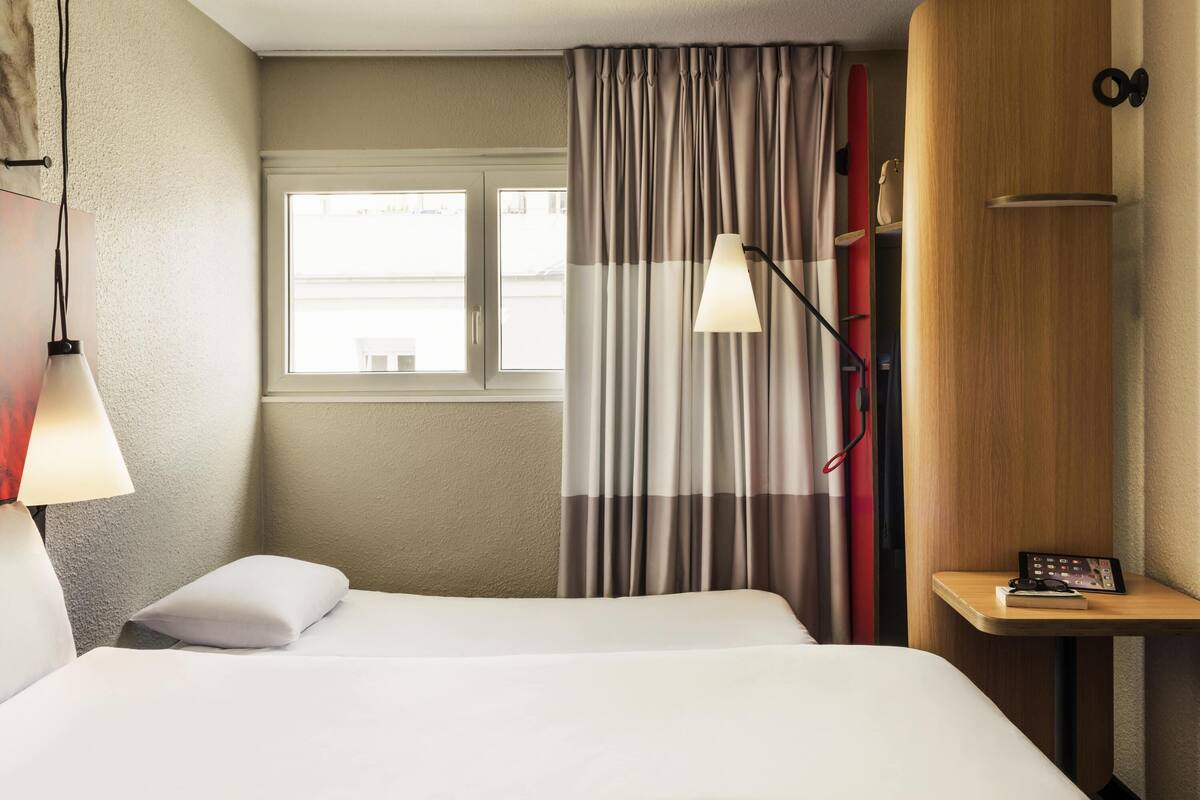 A hotel room with a bed, a desk with a lamp, a window, and curtains.