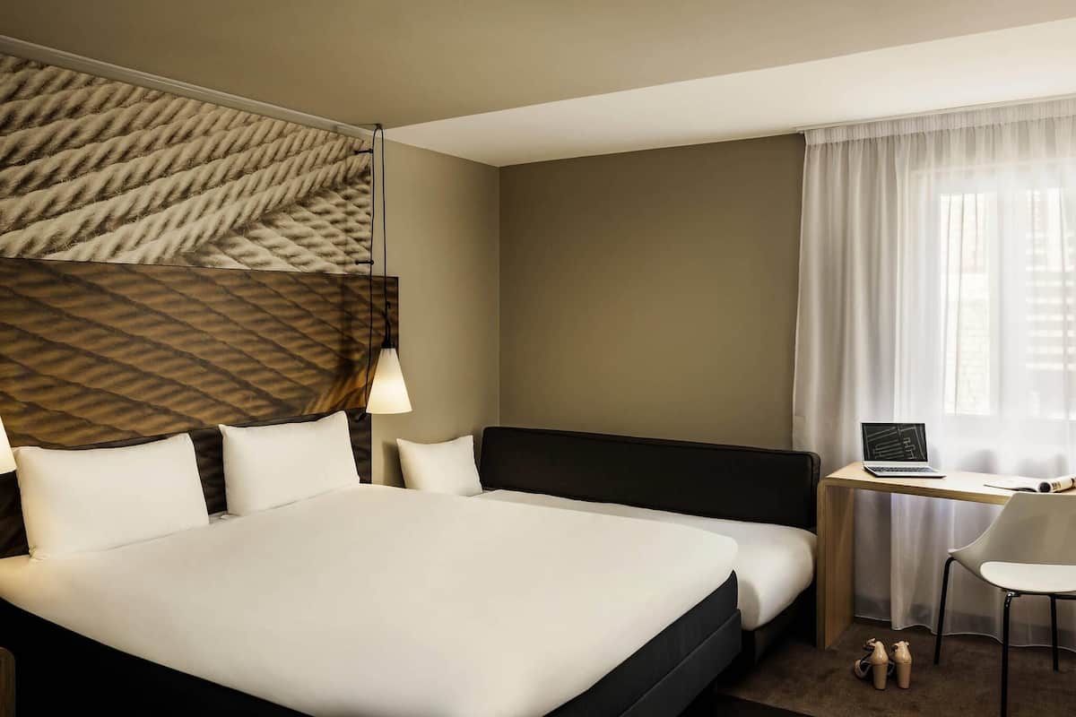 A hotel room with a large bed, a desk with a laptop, a chair, and a window with sheer curtains.