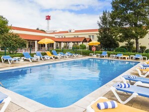 Una piscina con sillas de descanso y sombrillas, un edificio de techo rojo y una bandera.
