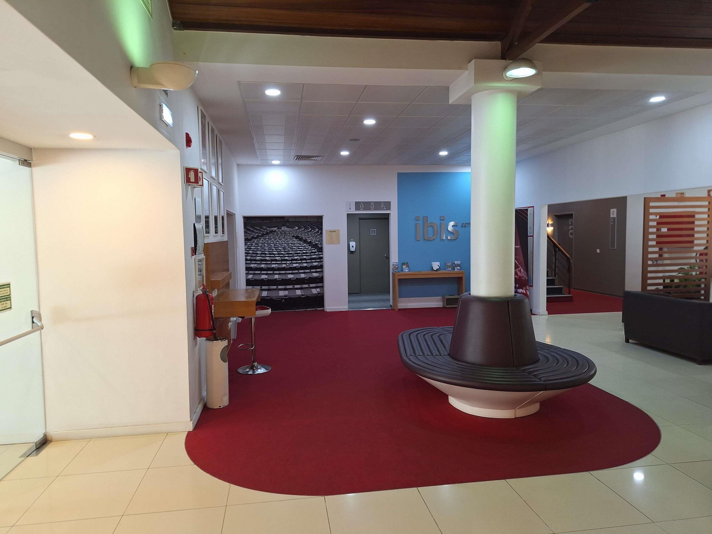 Un hall d’hôtel avec un tapis rouge, une colonne centrale, un comptoir d’accueil et un escalier.