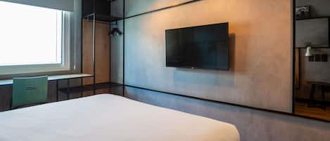A modern hotel room with a large bed, a flat-screen TV mounted on the wall, a desk with a chair, and a window providing natural light.