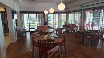 Een ruime eetruimte met meerdere tafels en stoelen, grote ramen met zonwering en hangende吊り灯.