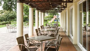 An outdoor patio with tables and chairs, a covered area with a wooden roof, and a playground in the background. - ibis Setubal (Setubal)