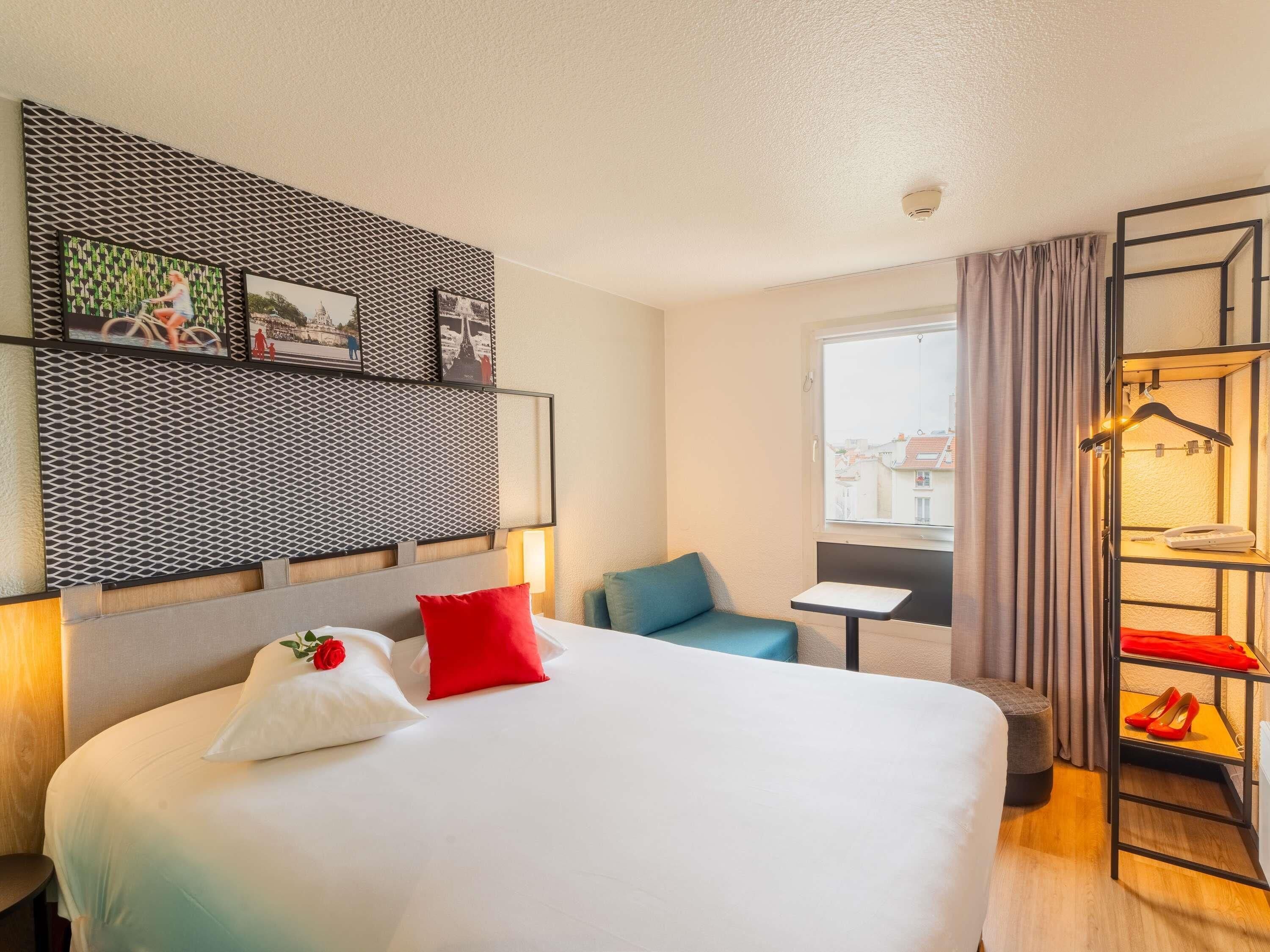 Une chambre d’hôtel avec un lit, une petite table, une chaise, une armoire avec des vêtements suspendus et une fenêtre donnant sur un paysage agréable.