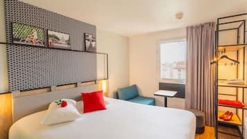 A hotel room with a bed, a small table, a chair, a wardrobe with hanging clothes, and a window with a view.