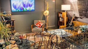Un área de estar moderna con un televisor de pantalla plana, un perchero de madera, un sillón estampado, una mesa de centro de vidrio y una estantería con libros y una guitarra.