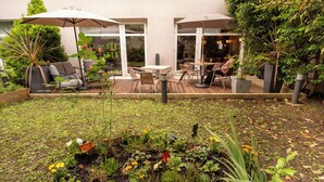 Un patio al aire libre con mesas y sillas, rodeado de plantas en macetas y un jardín.
