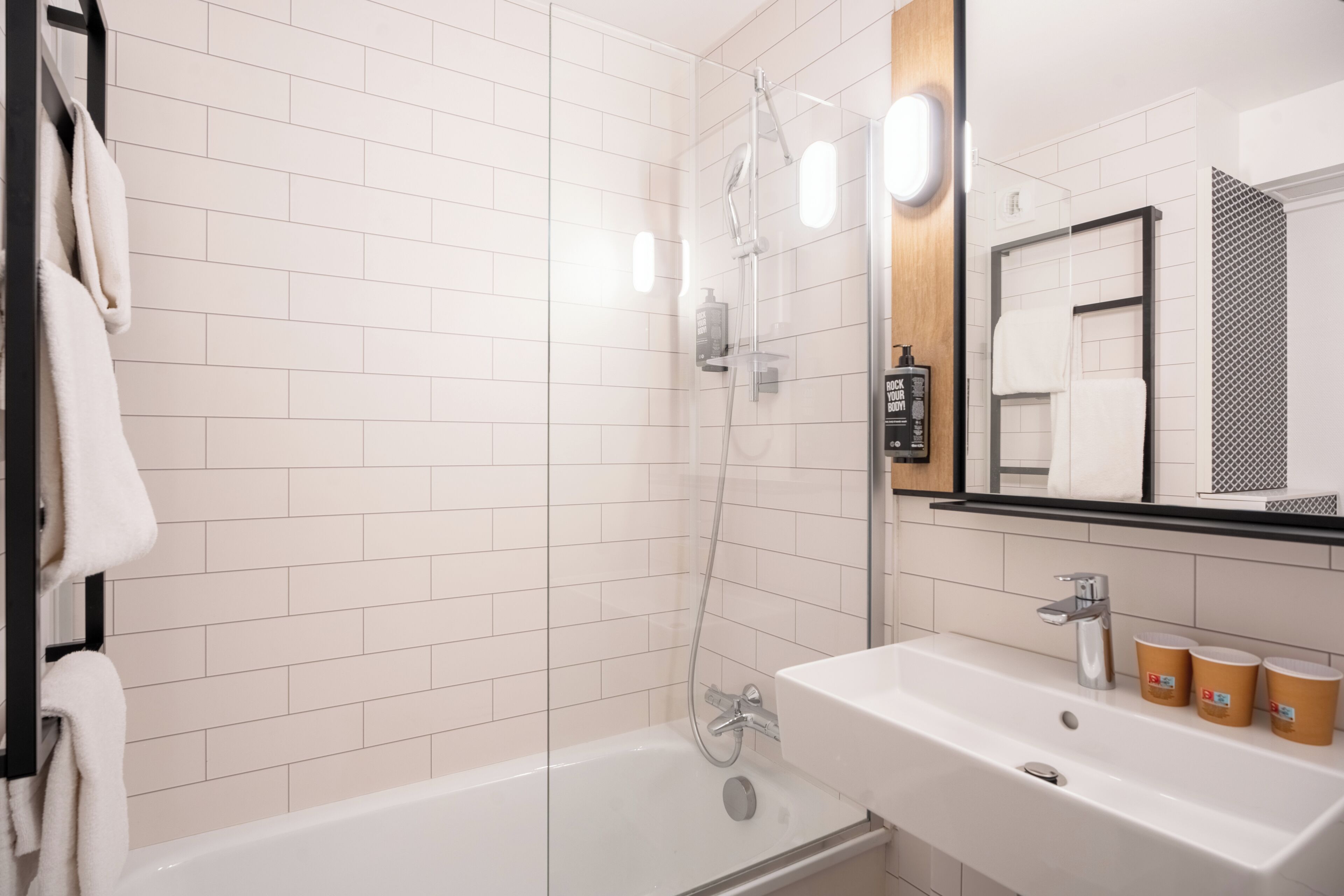 Un baño moderno con paredes de azulejos blancos, una ducha con cerramiento de vidrio, un lavabo blanco con dos vasos y un espejo con toallero.