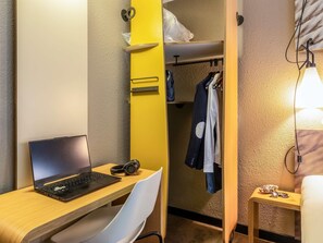 Una habitación pequeña con una computadora portátil sobre un escritorio, una silla, un perchero con ropa y una mesita de noche con una lámpara.