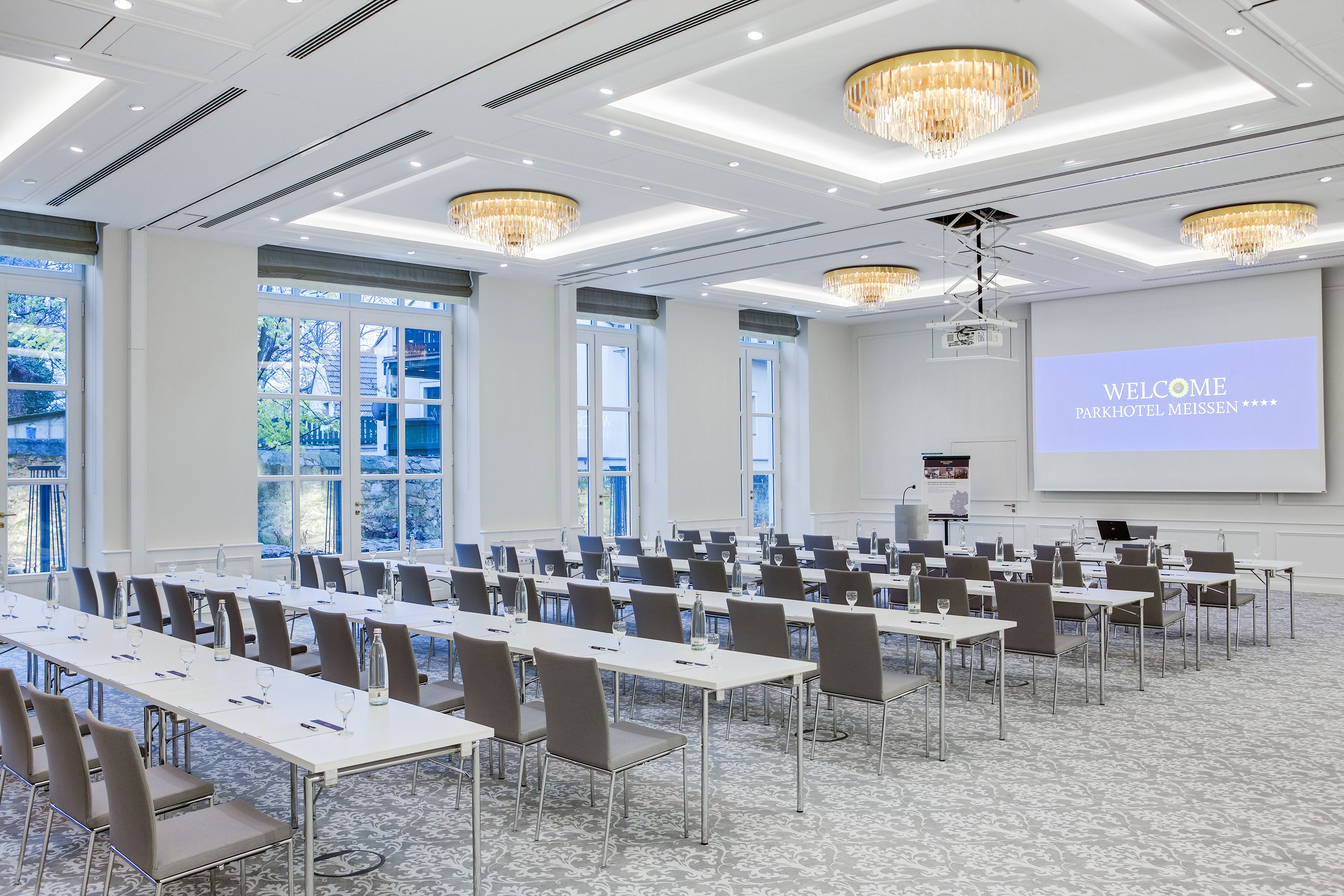 Ein Konferenzraum mit Reihen von Tischen und Stühlen, einer Projektionsleinwand mit der Aufschrift "WELCOME PARKHOTEL MEISSEN" und großen Fenstern für natürliches Licht.