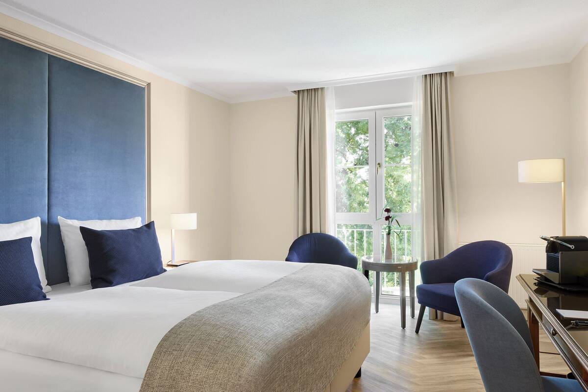 A hotel room with a large bed, two chairs, a small table, a desk with a computer, and a window with curtains.