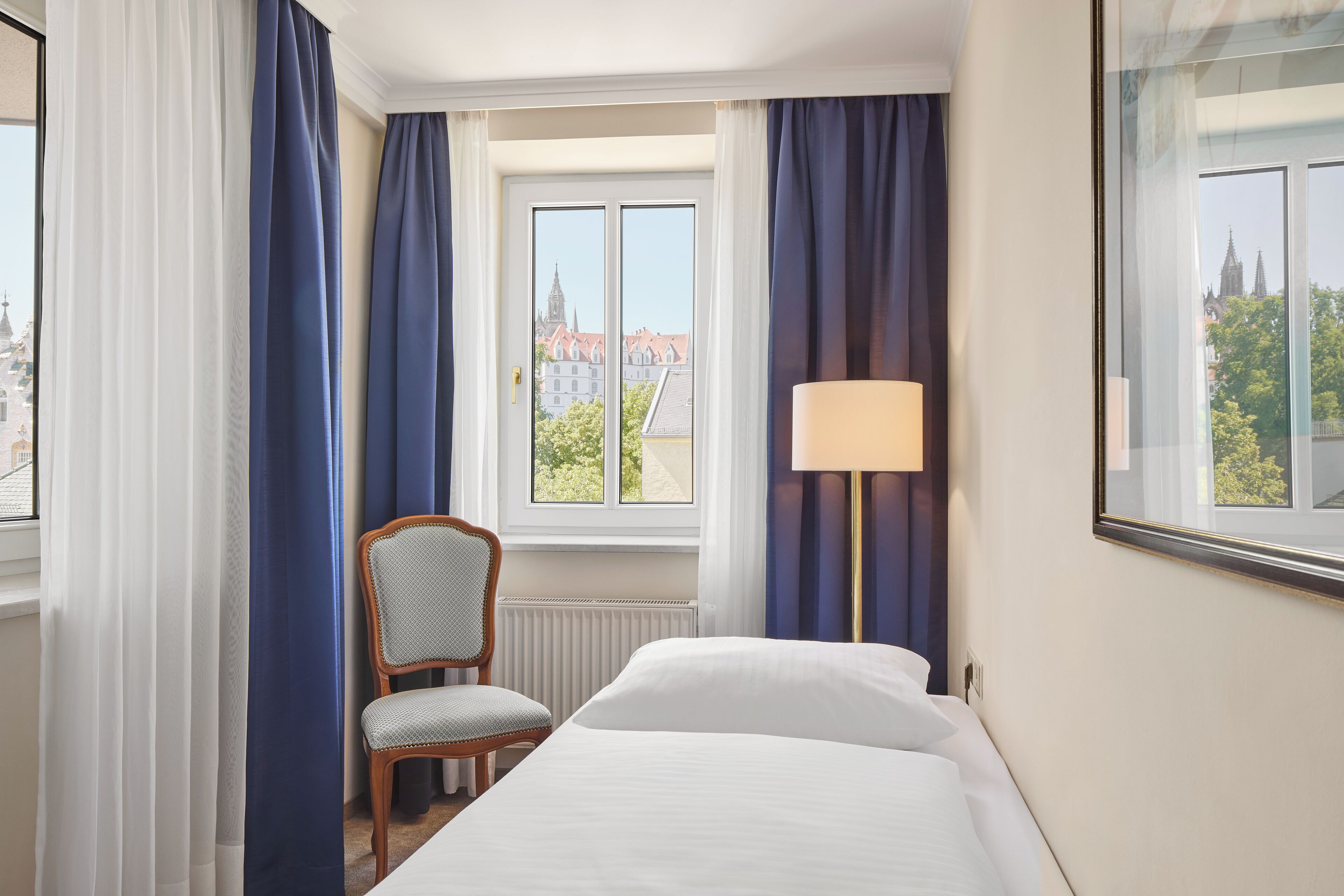A hotel room with a bed, a chair, a window with curtains, and a lamp.
