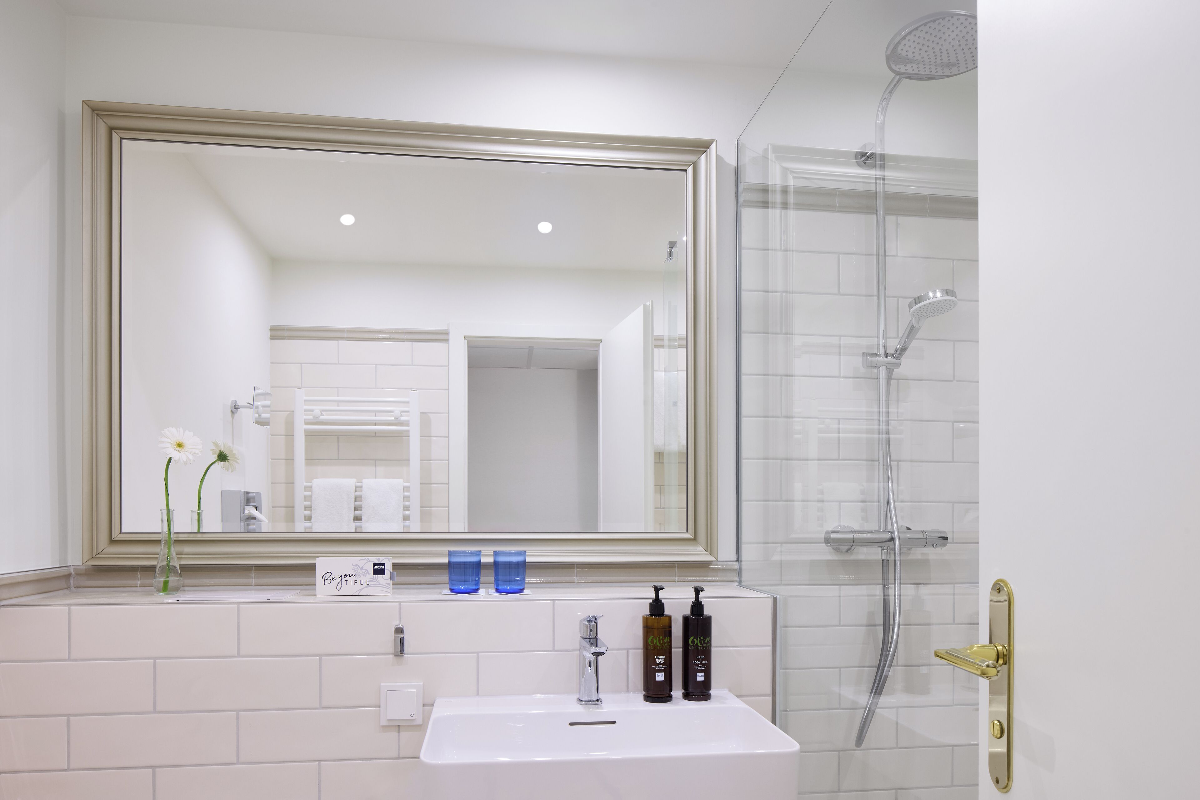 A bathroom with a large mirror, a sink, two bottles of shampoo, a vase with flowers, and a shower area.