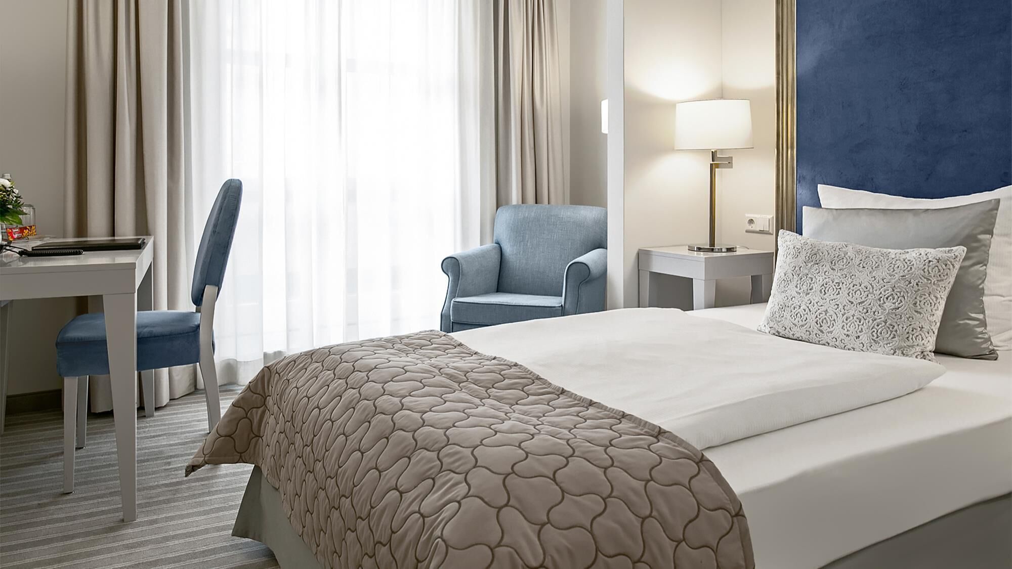 A hotel room with a bed, a desk, a chair, a lamp, and a nightstand.