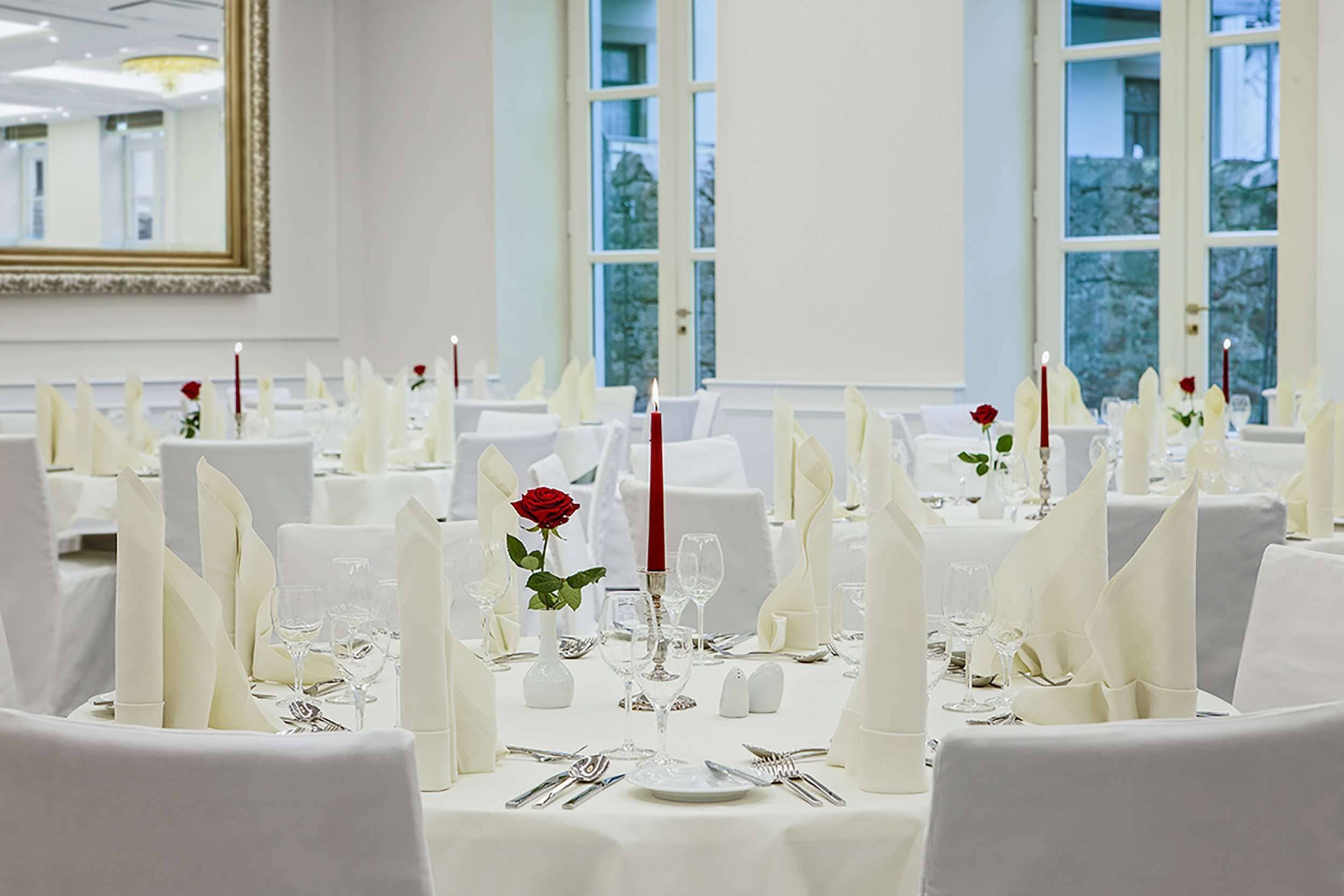 A banquet hall with round tables set for a formal event, featuring white tablecloths, white napkins, and red candles.