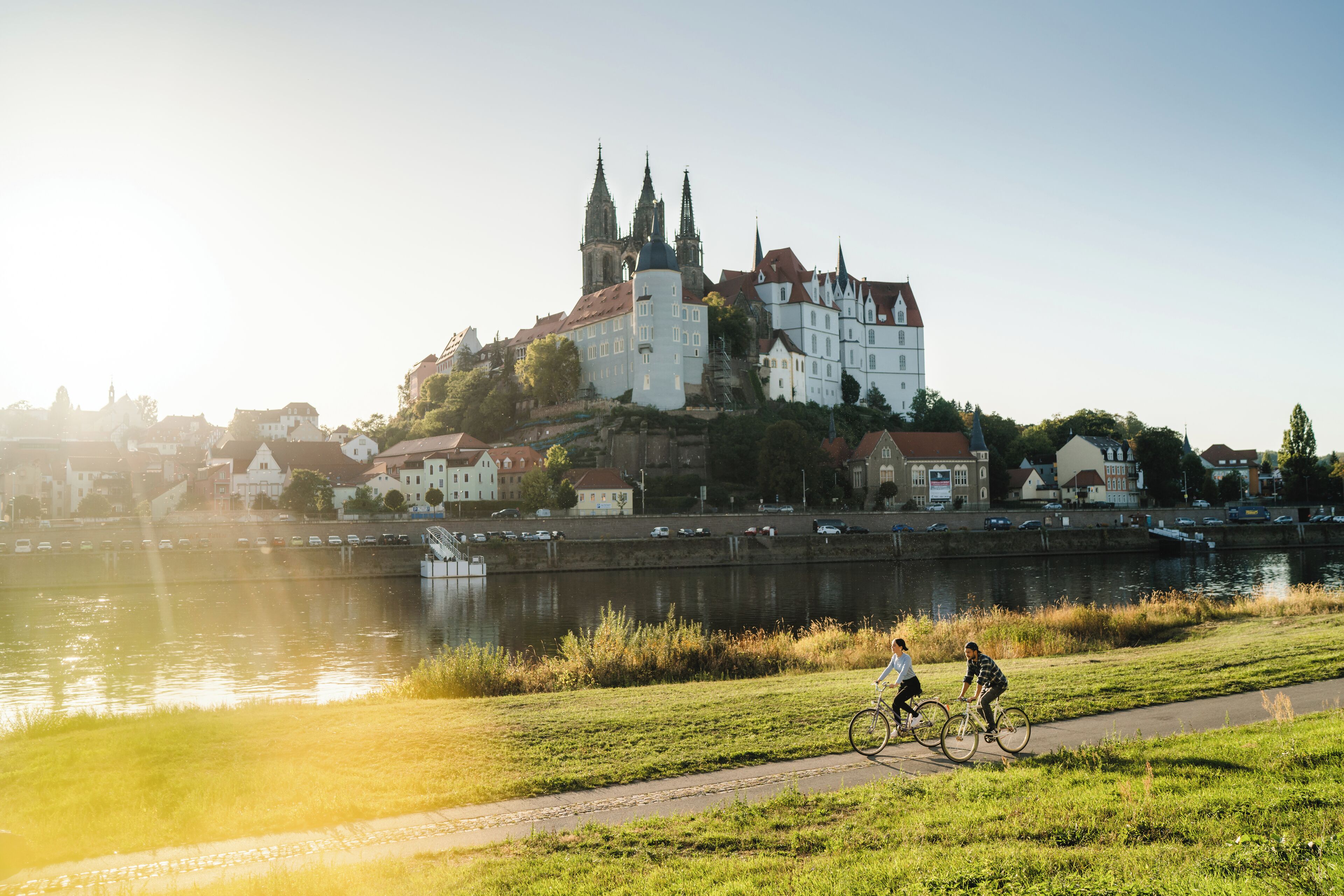 A riverside view with a historic castle and a paved path where two cyclists are riding.